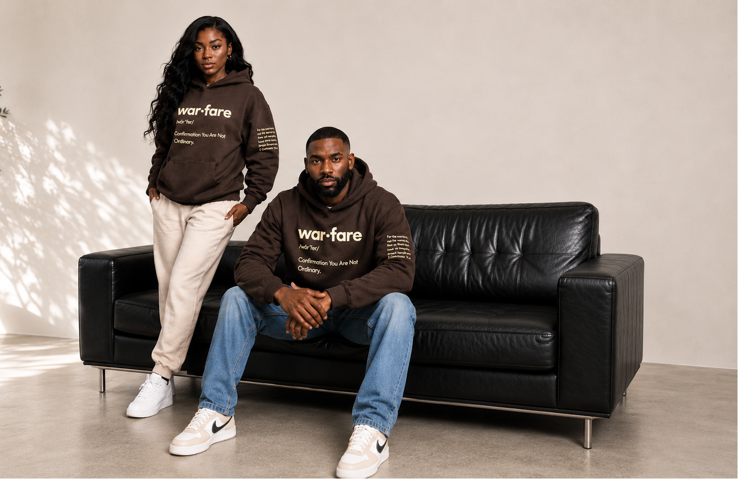 Two people wearing 'warfare' branded hoodies sitting on a black leather sofa in a modern room.