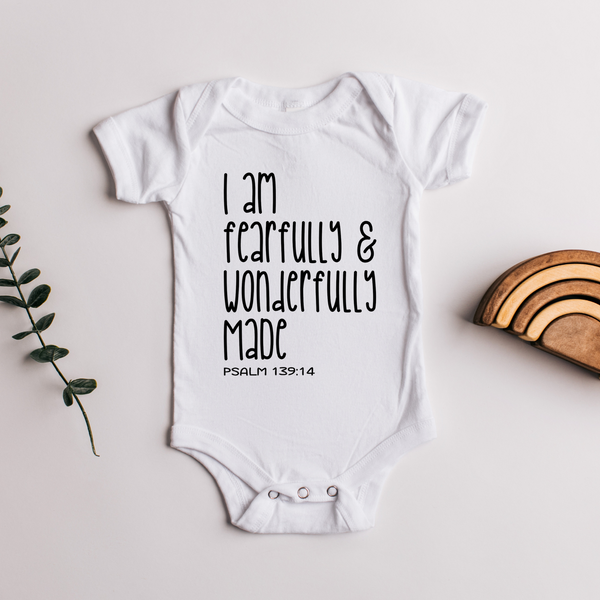 White baby onesie featuring the scripture phrase “I am fearfully & wonderfully made - Psalm 139:14” in bold black text. Flat lay styled with a eucalyptus sprig and a wooden rainbow toy, ideal for Christian newborn photography and faith-based baby gifts.