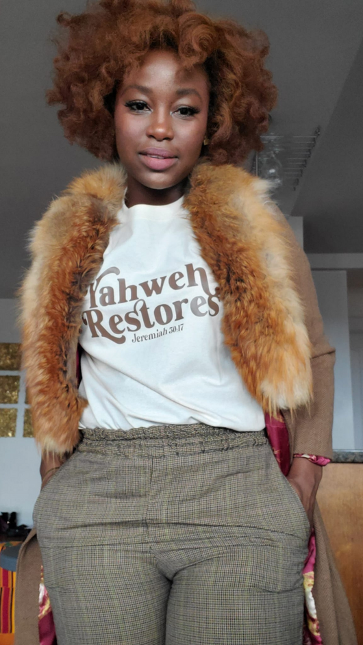 Confident African American woman wearing a white "Yahweh Restores" Christian T-shirt with brown lettering, styled with plaid pants and a tan fur-collared coat. Inspired by Jeremiah 30:17, this faith-based apparel captures strength, restoration, and vintage-inspired fashion.